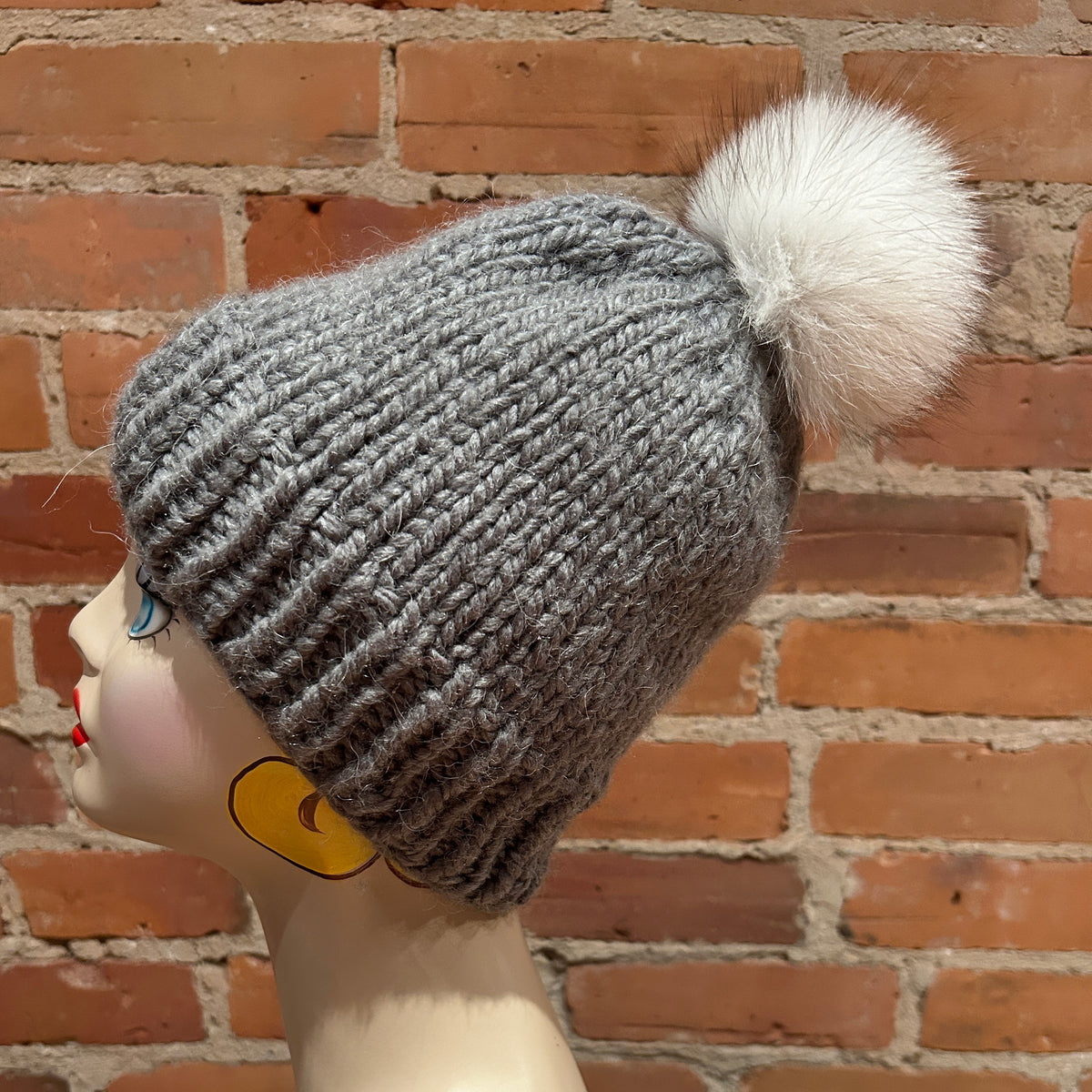 A gray ribbed knit beanie hat with a fluffy white pom-pom, displayed on a mannequin head against a red brick wall background.
