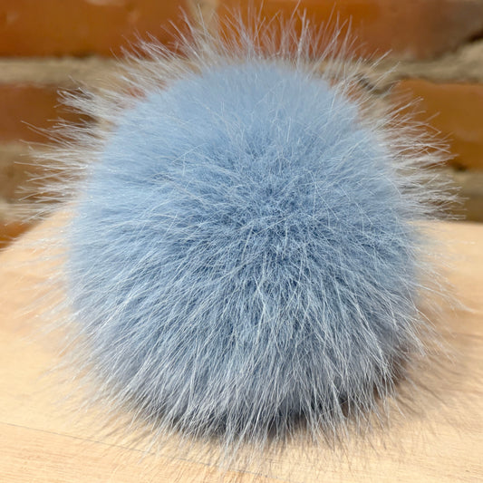 A fluffy, round pom-pom accessory in a dusty light blue-gray color, resting on a light wood surface with a blurred brick background.