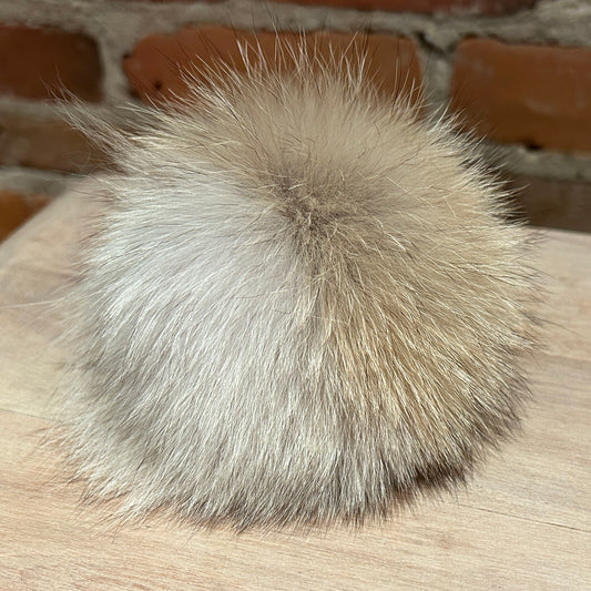 A fluffy coyote fur pom-pom ball in a light beige and brown color, resting on a light wood surface with a brick wall background.