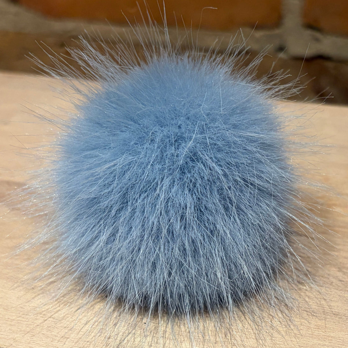 A fluffy, light blue faux-fur pom-pom accessory resting on a light-colored wooden surface with a blurred brick wall in the background.