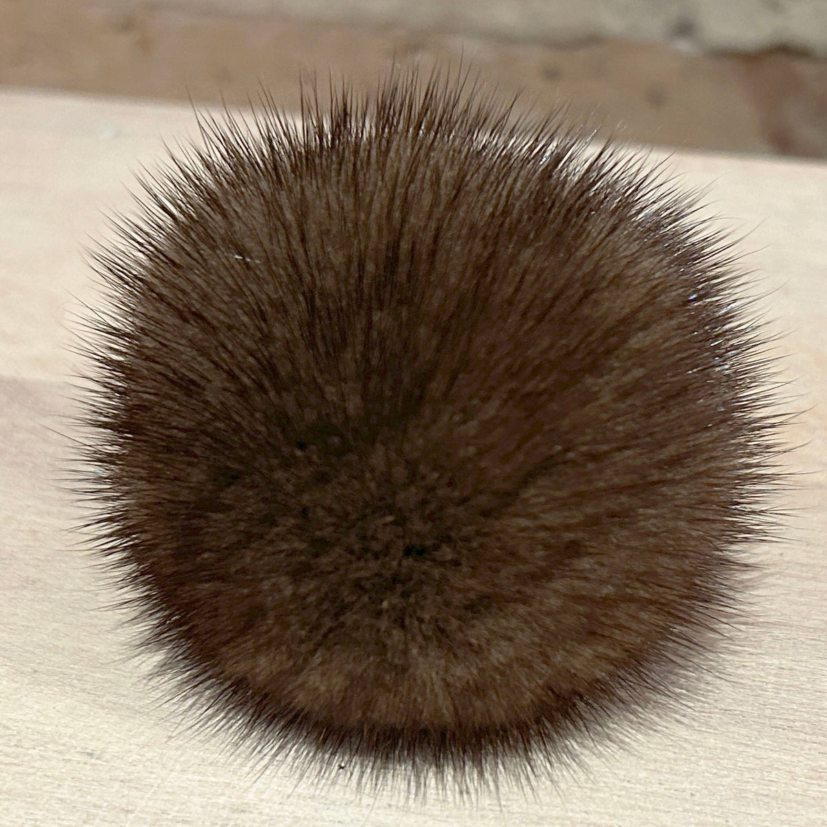 A round, fluffy pom-pom in a dark brown color, resting on a light-colored wooden surface with a blurred brick wall in the background.