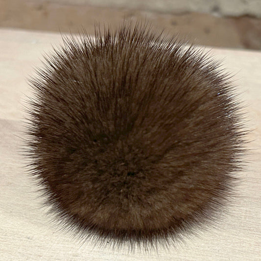 A round, fluffy pom-pom in a dark brown color, resting on a light-colored wooden surface with a blurred brick wall in the background.