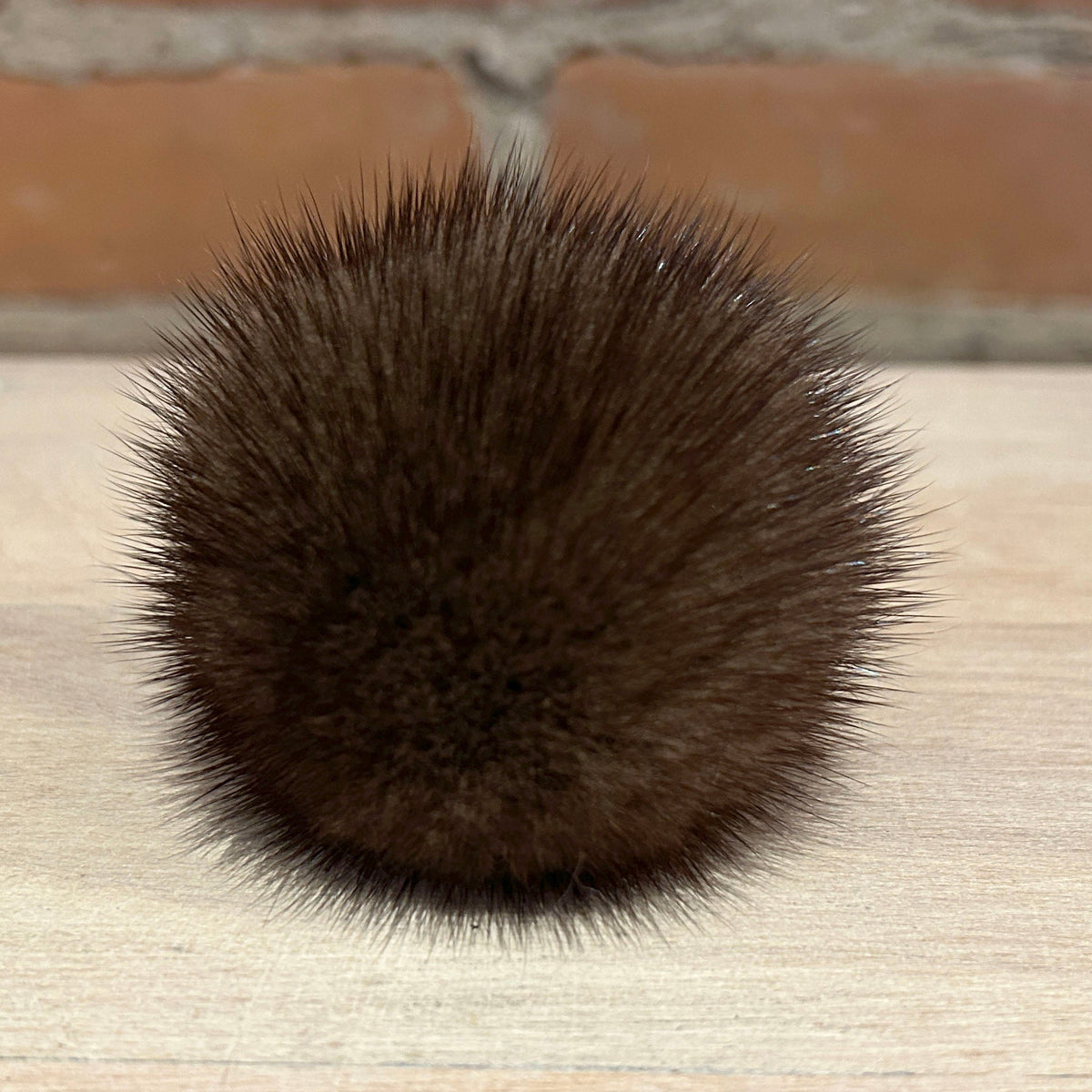 A round, dark brown mink fur pom-pom accessory with dense, spiky fibers, resting on a light wood surface with a blurred brick wall background.