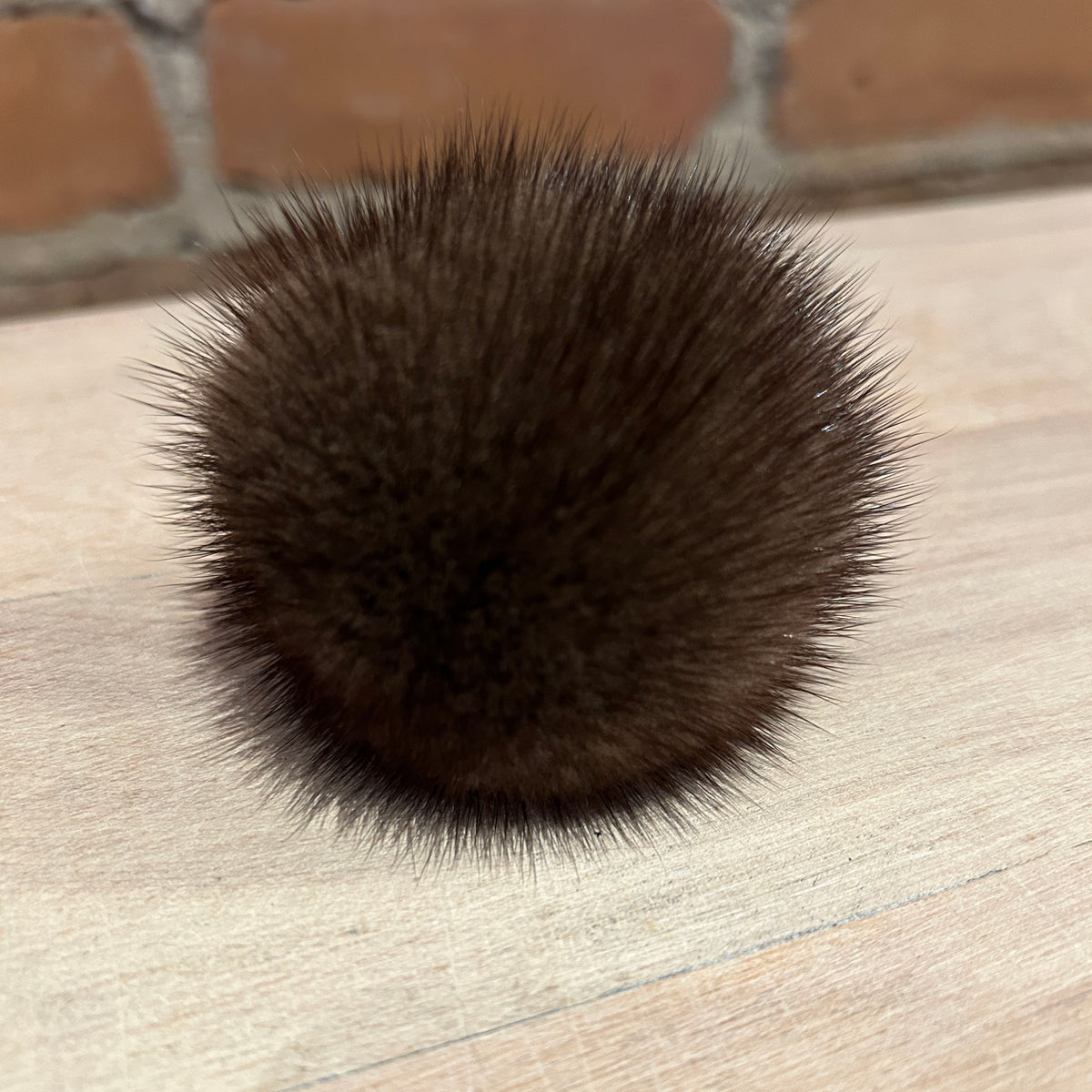 A dark brown mink fur pom-pom accessory resting on a light-colored wooden surface with a blurred brick wall in the background.