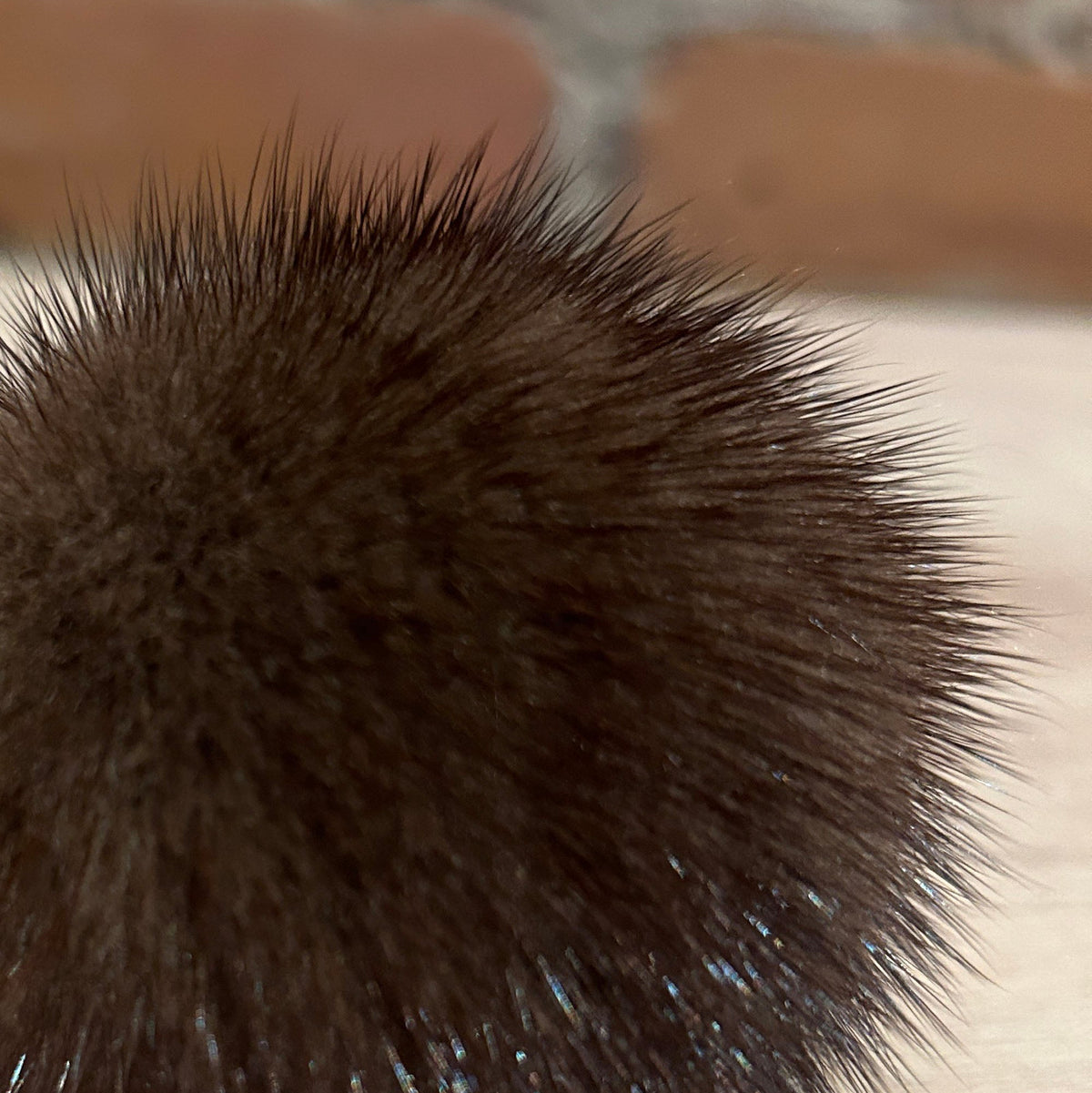 A close-up of a dark brown mink fur hat pom pom resting on a light wood surface with a blurred brick wall in the background.