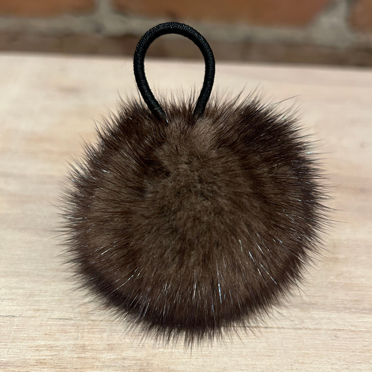 A dark brown mink fur pom-pom with a black elastic hair tie, resting on a light wood surface.