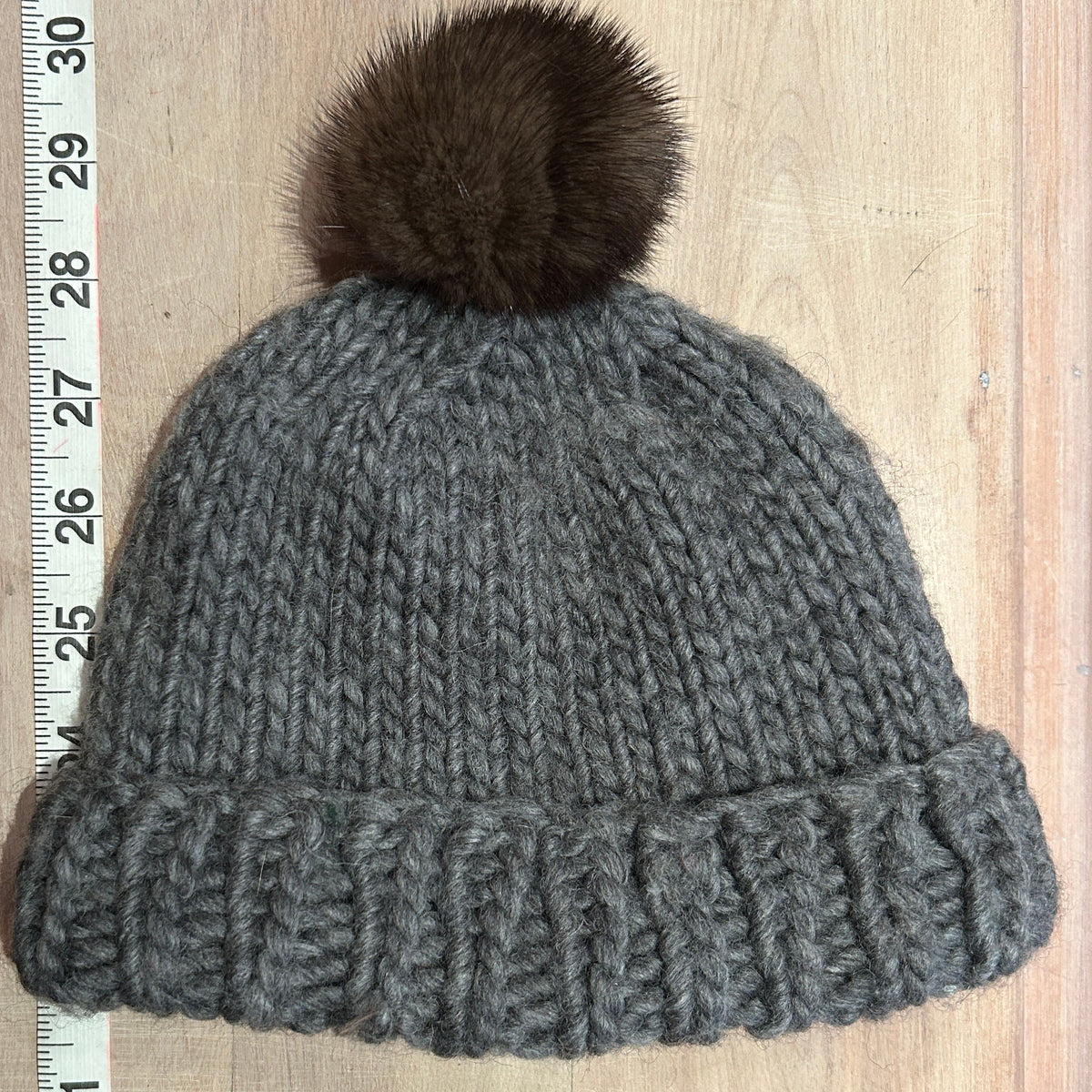 A gray chunky knit beanie with a dark brown mink fur pom-pom, laid flat on a light wood surface next to a white measuring tape.