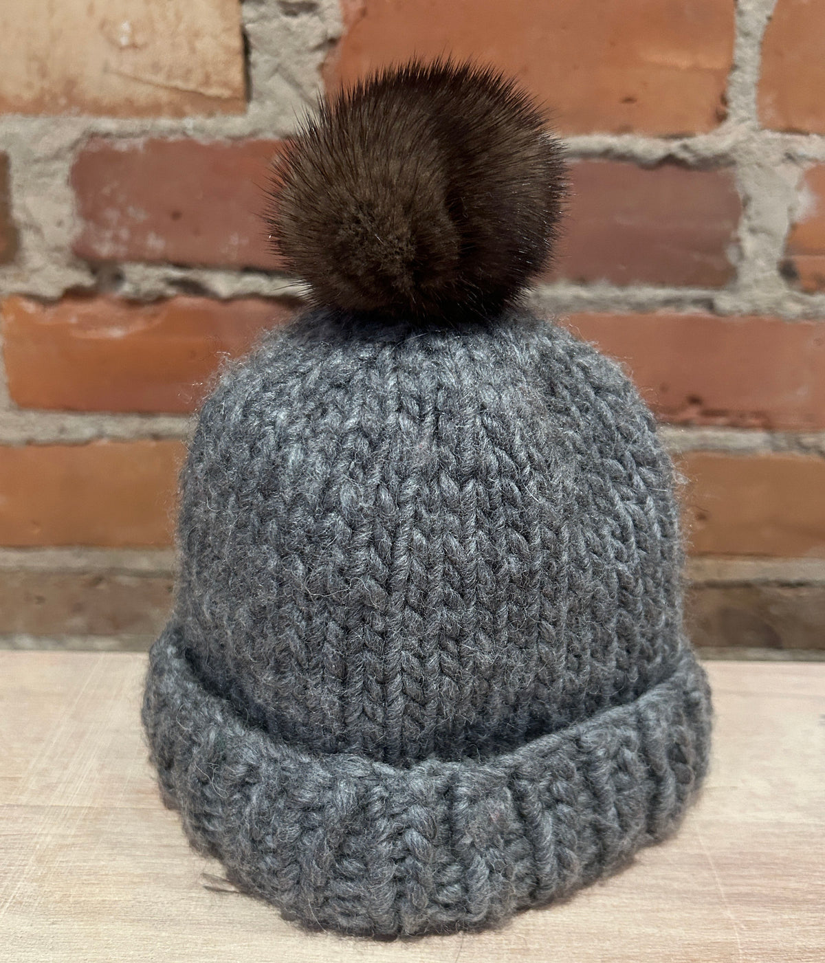 A gray chunky knit beanie hat with a dark brown mink  fur pom-pom, displayed on a light wood surface against a rustic red brick wall background.