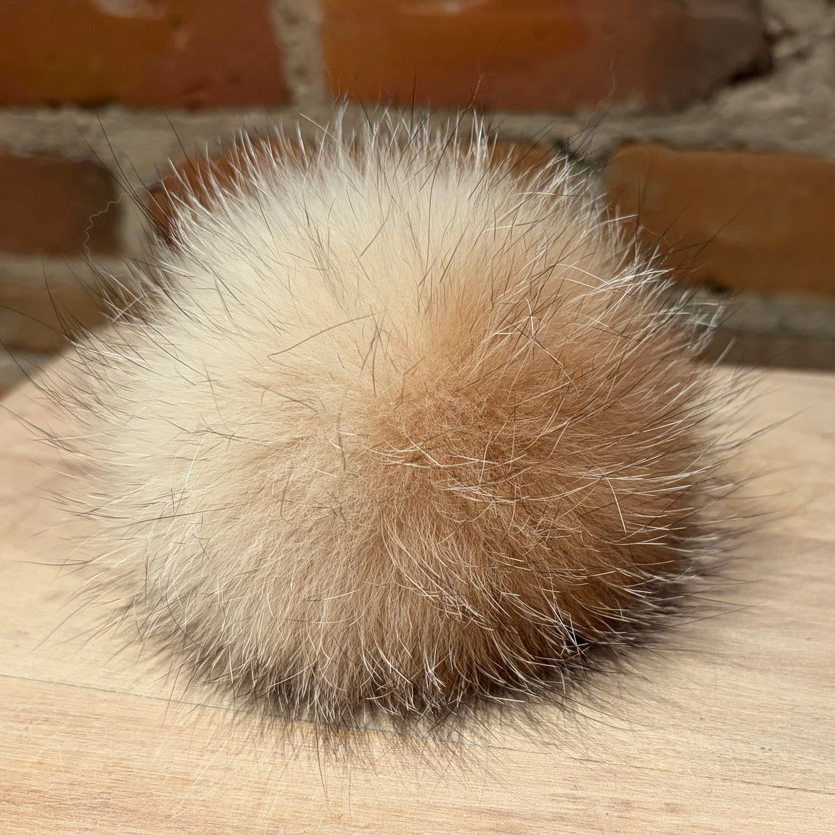 A fluffy fox-fur pom-pom ball in a natural beige and light brown color, resting on a light wood surface with a blurred brick wall background.