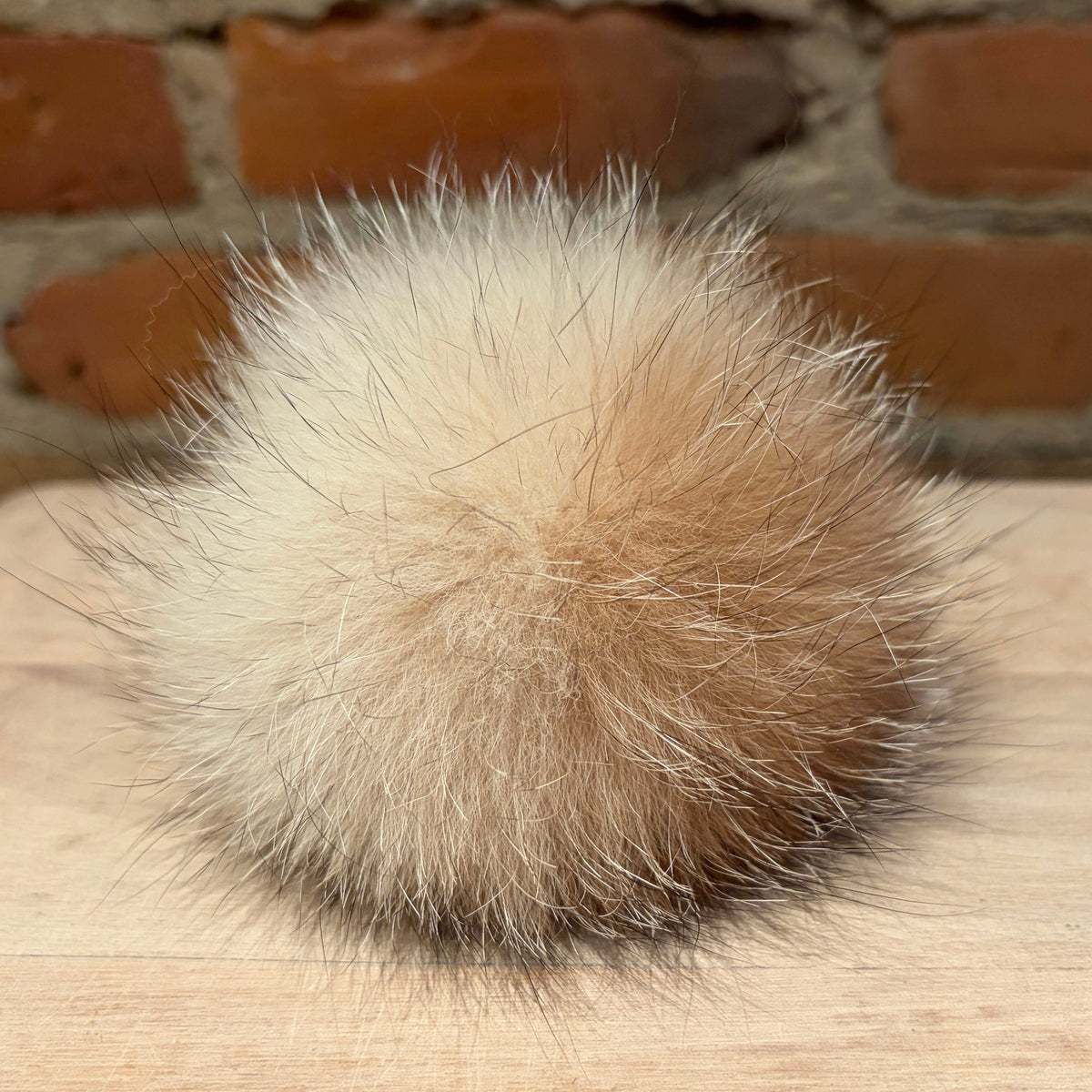 A fluffy fox fur pom-pom ball in a natural beige and light brown color, resting on a light wooden surface with a blurred brick wall background.