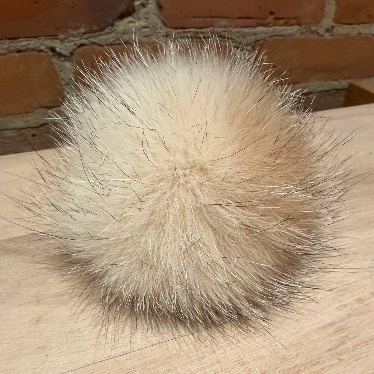 A fluffy fox-fur pom-pom ball in a light cream and beige color, resting on a light wood surface with a brick wall background.
