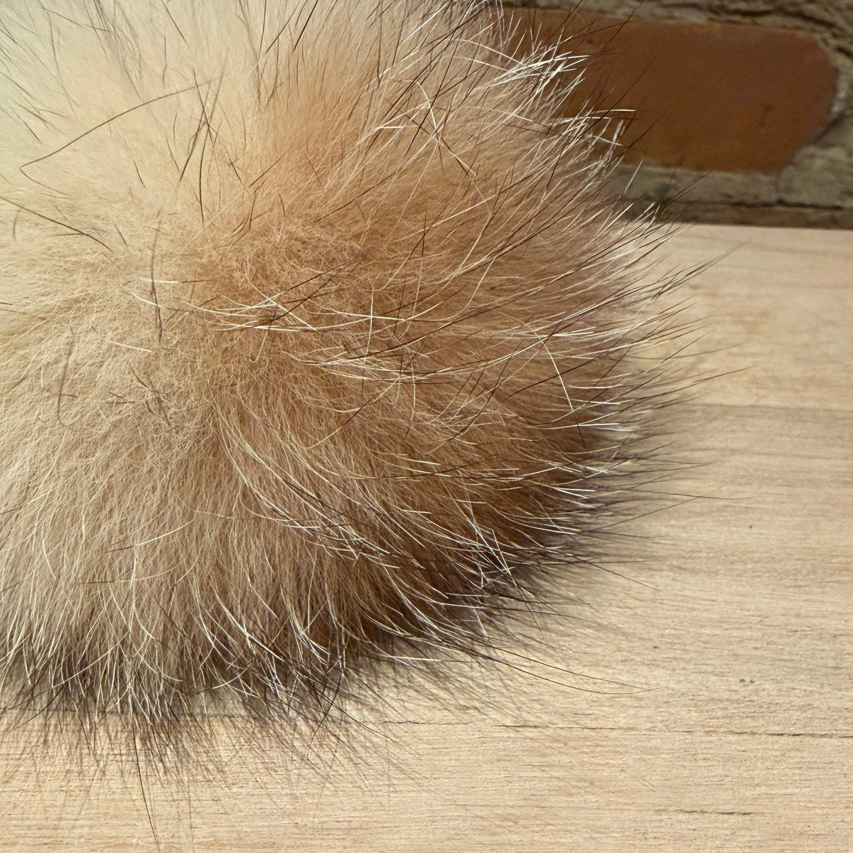 A close-up of a fluffy fox fur pom-pom accessory in light tan and cream tones, resting on a light wood surface with a brick wall background.