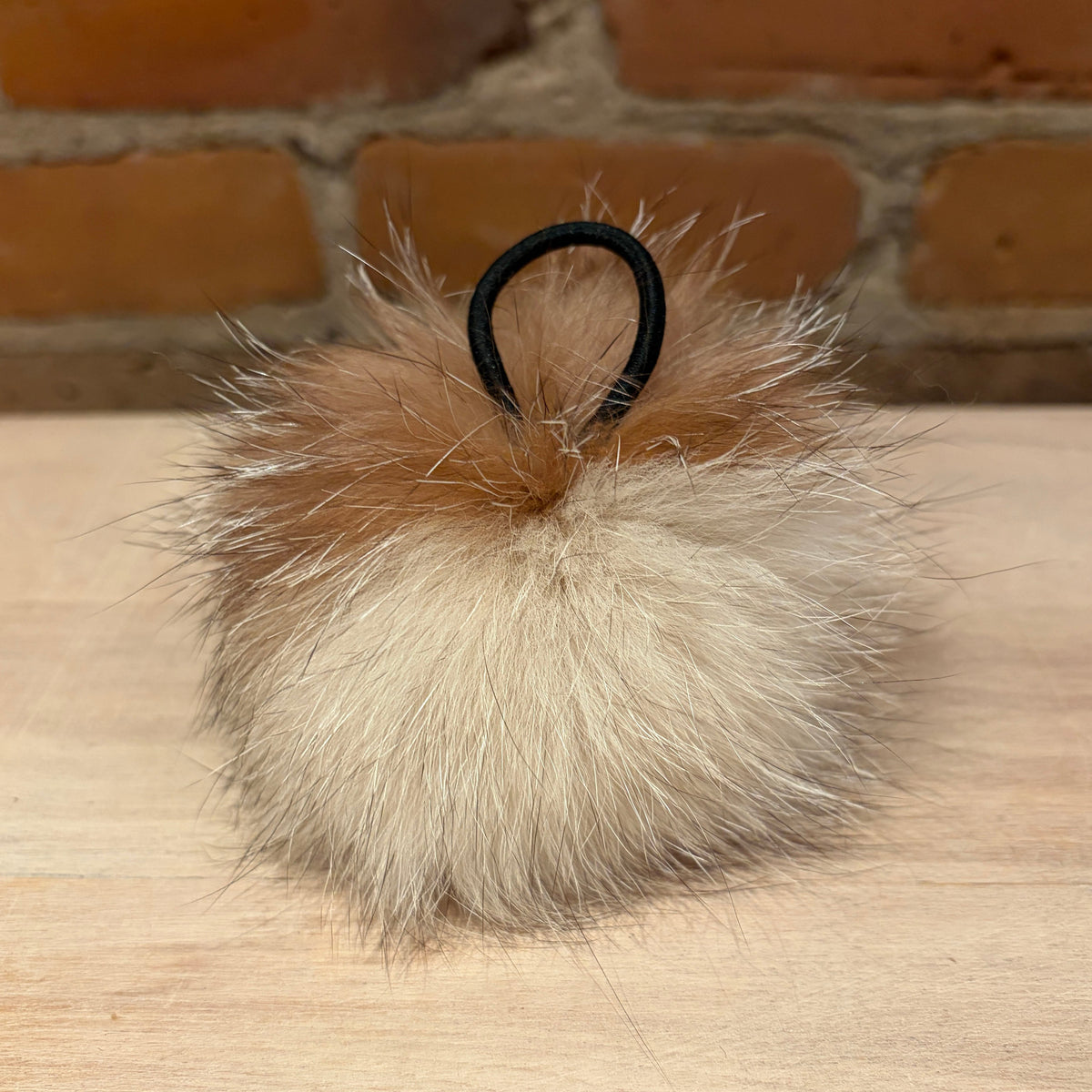 A fluffy fox-fur pom-pom hair accessory in beige and light brown tones with a black elastic loop, resting on a light wood surface with a brick wall background.