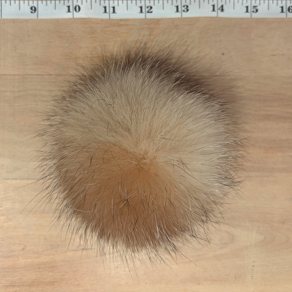 A fluffy fox-fur pom-pom ball in shades of beige, tan, and brown, shown from above on a light wood surface with a measuring ruler at the top for scale.