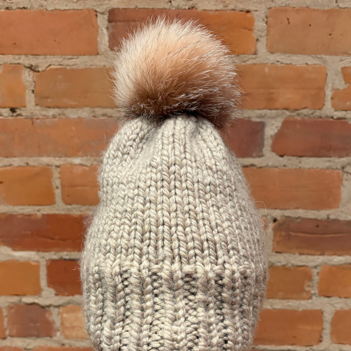 A chunky knit beanie hat in a light oatmeal gray color with a large tan foxfur pom-pom, displayed on a mannequin head against a brick wall background.