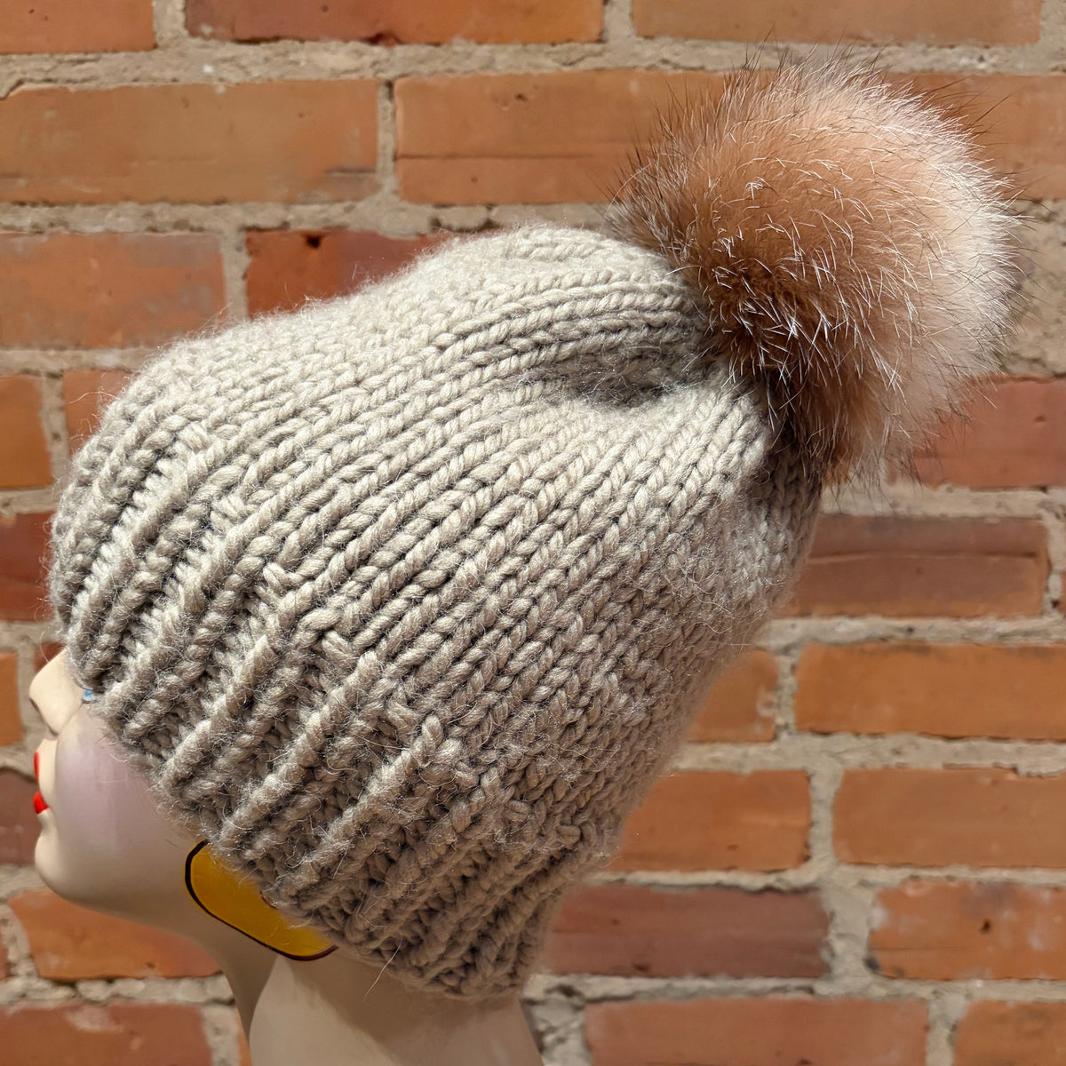 A light beige ribbed knit beanie with a fluffy brown and cream fox fur pom-pom, displayed on a mannequin head against a red brick wall background.