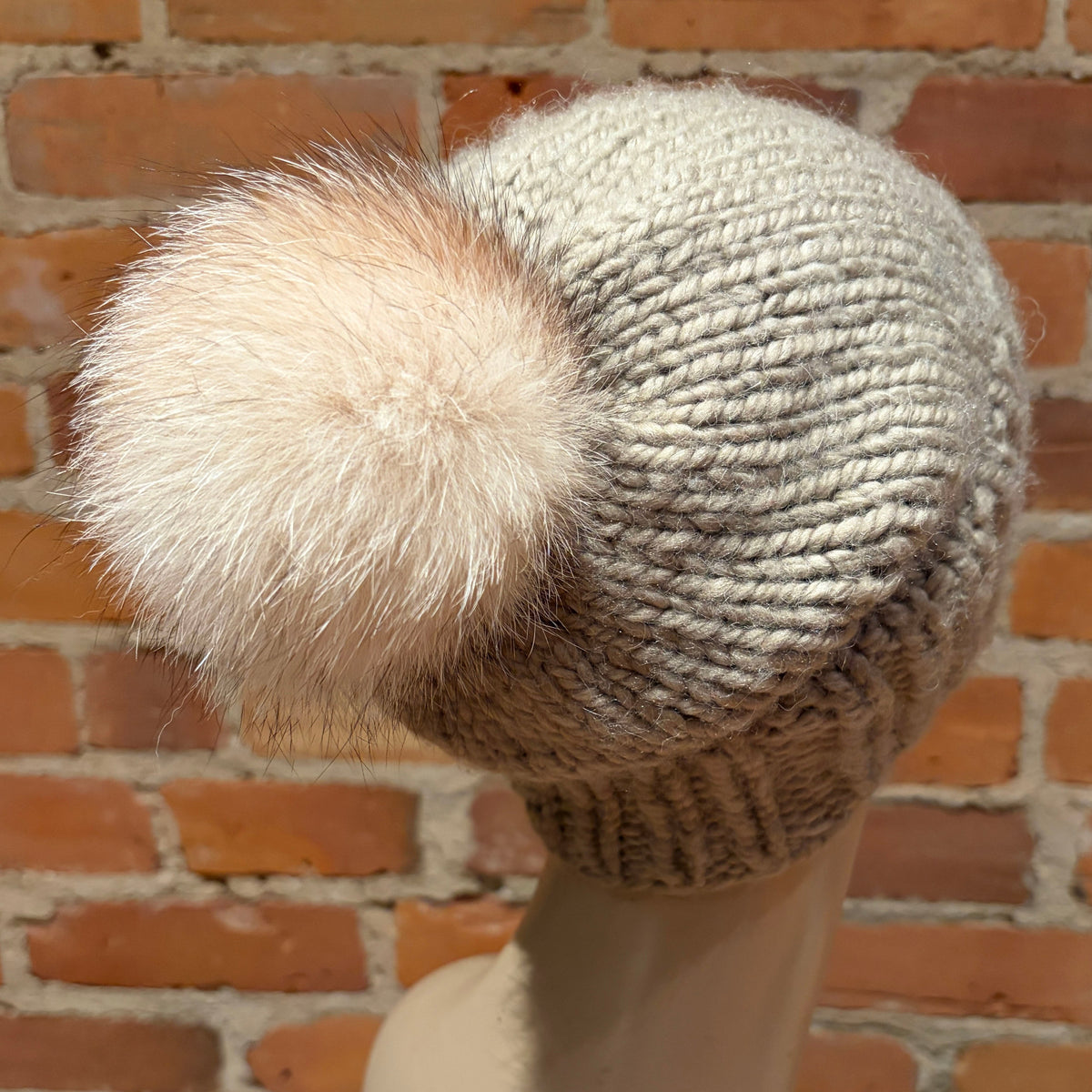 A chunky knit beanie hat in a light oatmeal gray color with a large fluffy cream fox fur pom-pom, displayed on a mannequin head against a brick wall background.