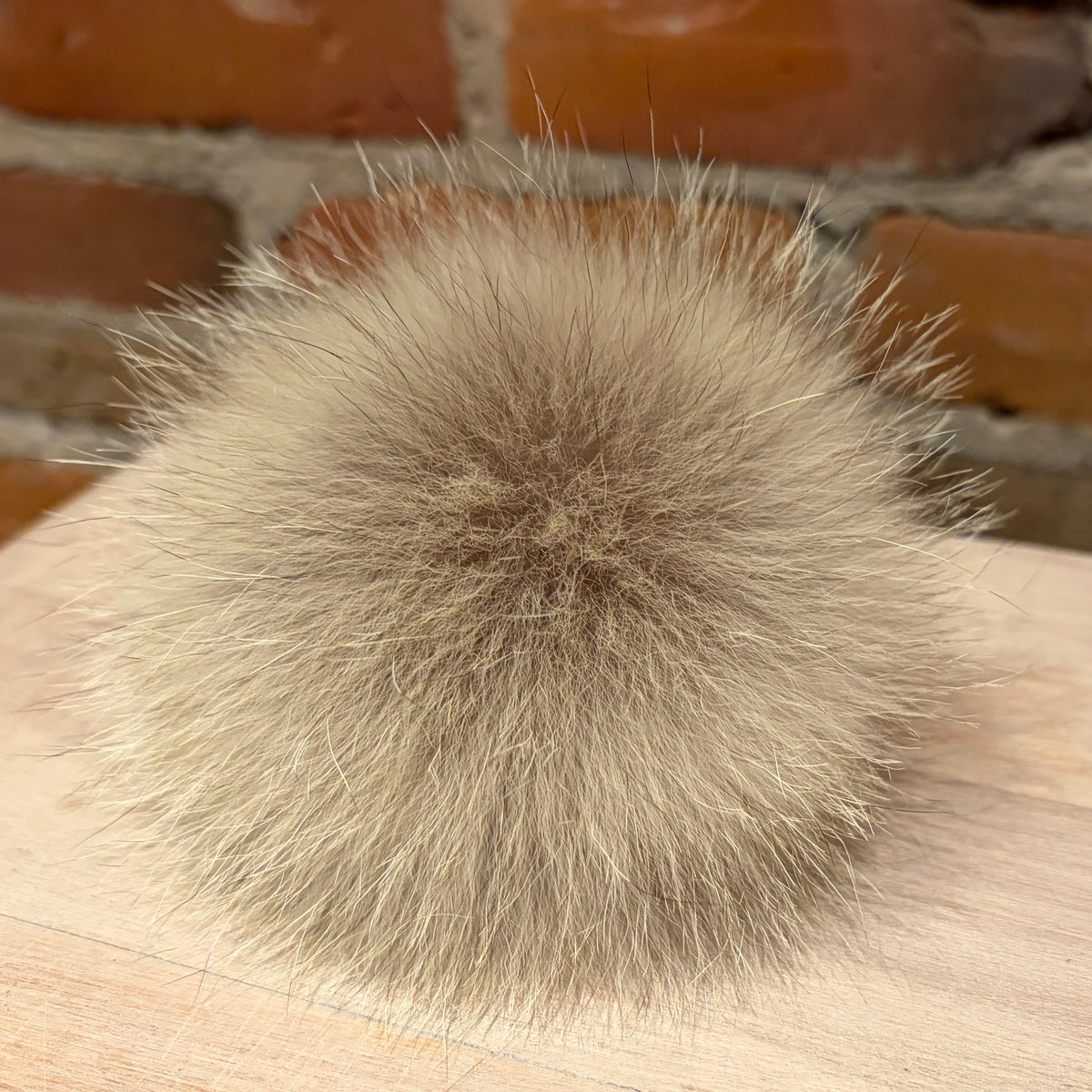A fluffy coyote fur pom-pom accessory in a light beige and brown color, resting on a light wood surface with a blurred brick wall background.