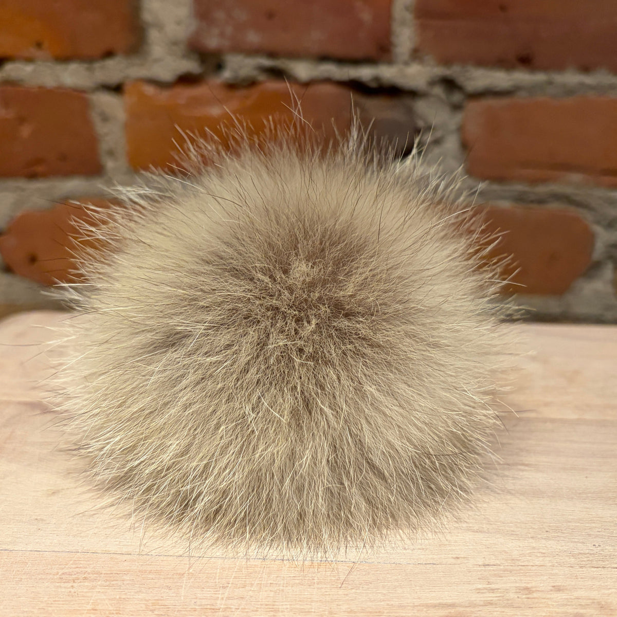 A round, fluffy coyote fur pom-pom in a light taupe-gray color, resting on a light wood surface with a brick wall background.