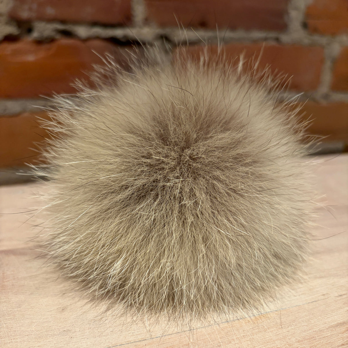 A fluffy coyote-fur pom-pom ball in a light taupe-gray color, resting on a light wood surface with a blurred red brick wall in the background.
