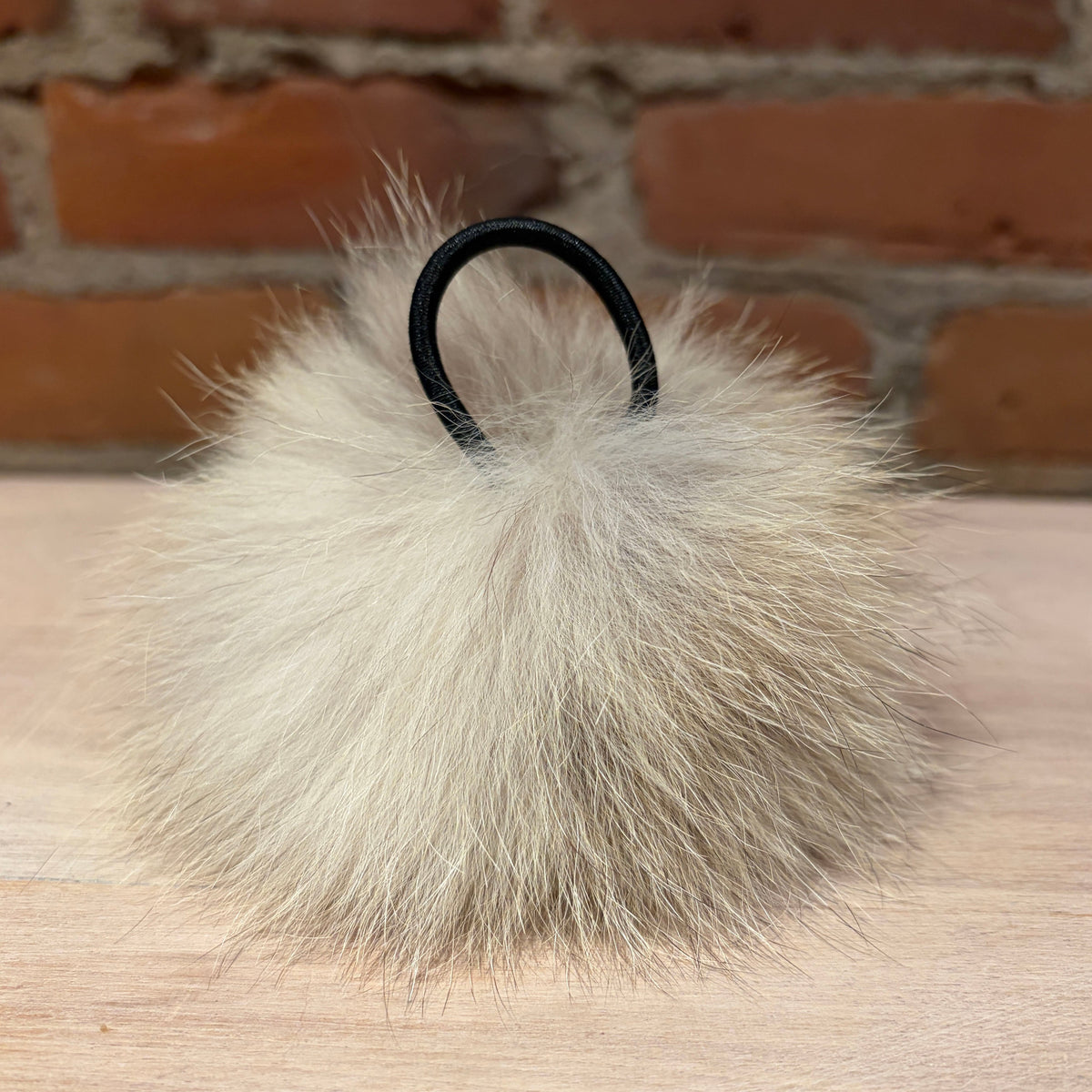 A fluffy pom-pom hat pom pom, in a light beige/cream coyote fur material with a black elastic loop, resting on a light wood surface with a blurred brick wall background.