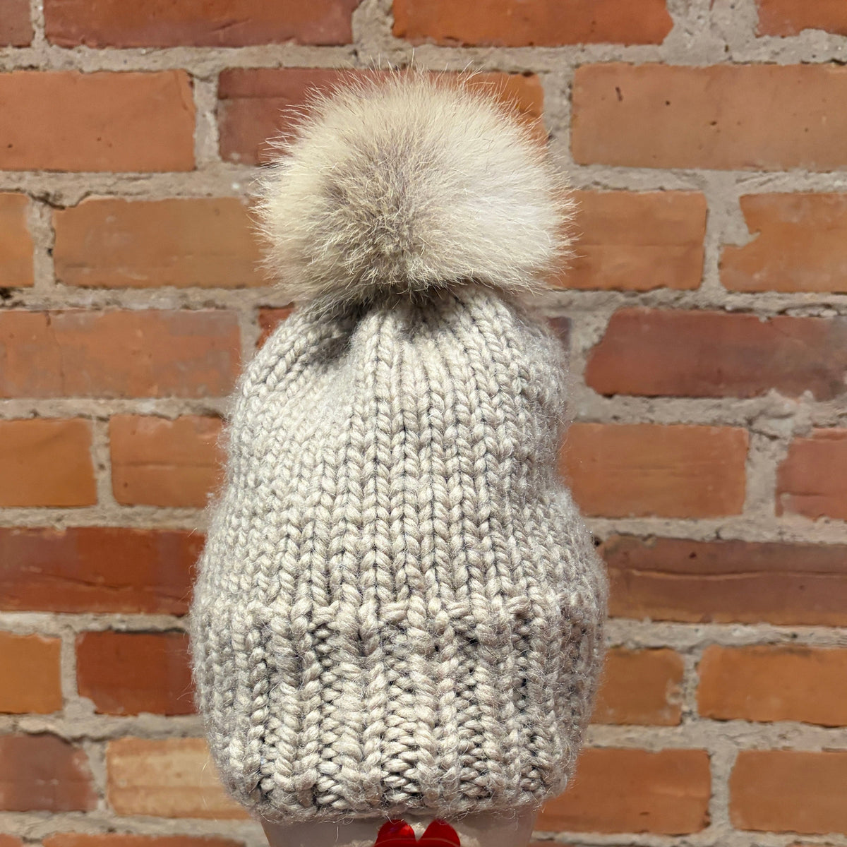 A chunky knit beanie in a light gray and white heathered yarn, topped with a large fluffy coyote-fur pom-pom, displayed on a mannequin head against a red brick wall background.