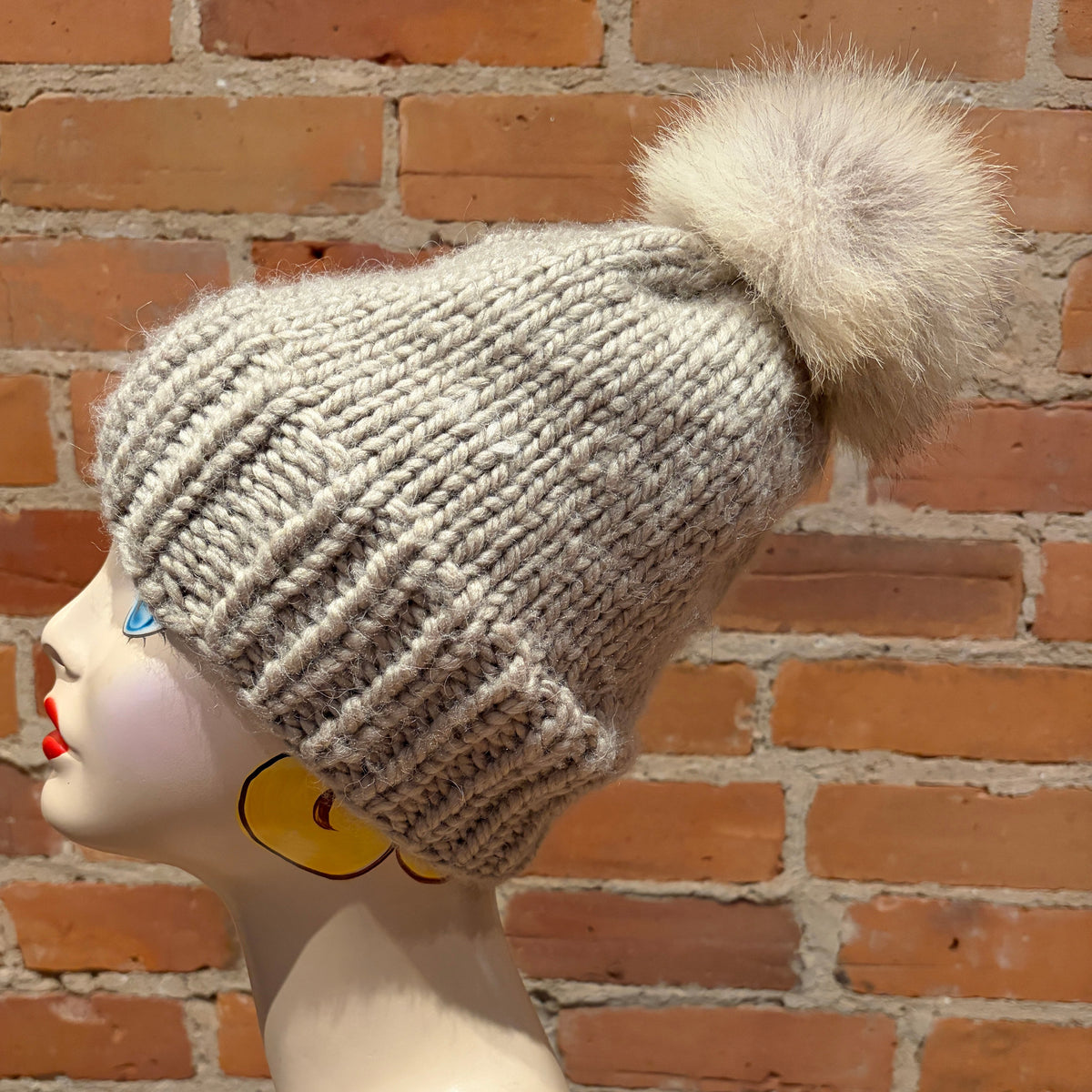 A light gray chunky knit beanie with a large cream coyote-fur pom-pom, displayed on a mannequin head against a red-brown brick wall background.