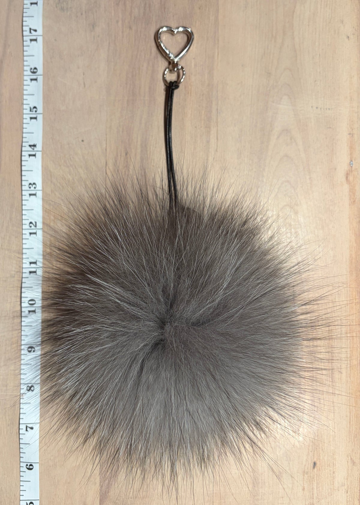 A gray silver fox fur pom-pom keychain or bag charm with a black strap and a silver heart-shaped metal ring, displayed on a light wood background next to a measuring ruler.