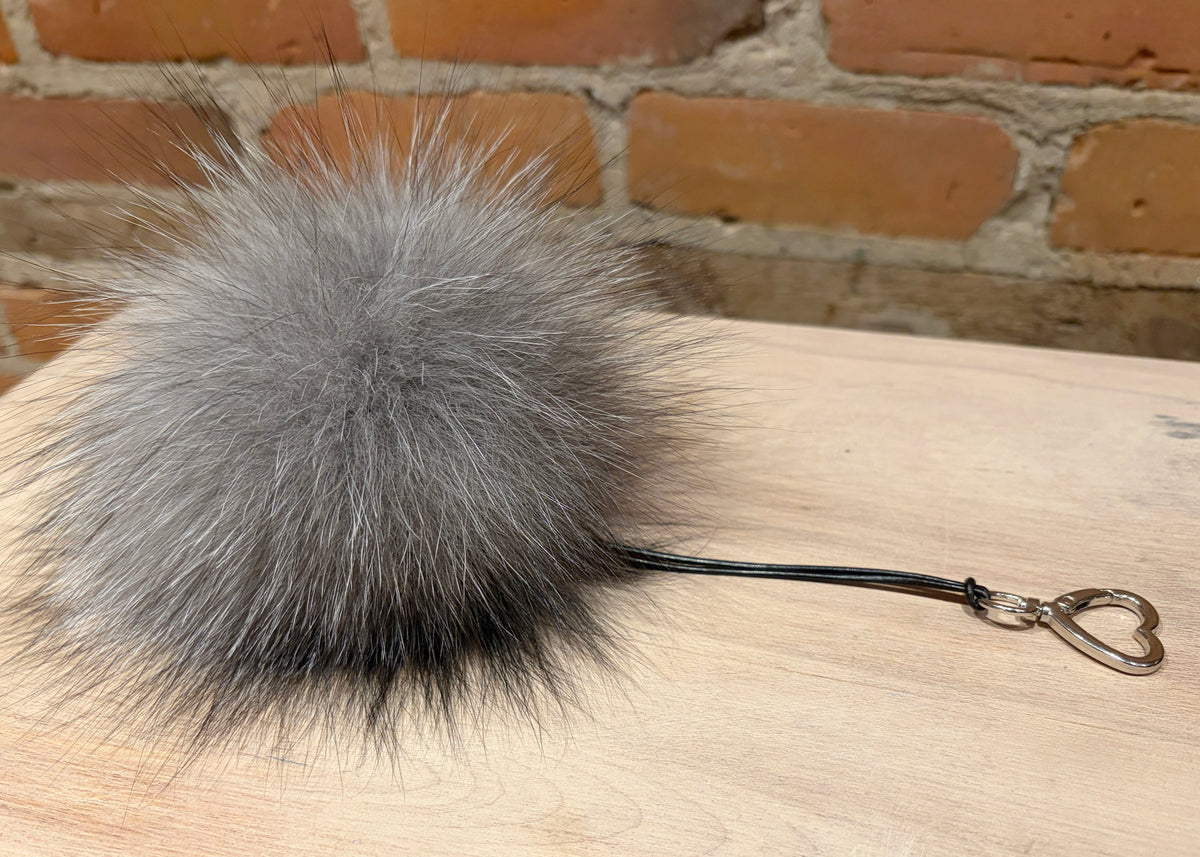 A fluffy gray faux fur pom-pom keychain with a black cord and silver heart-shaped keyring, resting on a light wood surface with a brick wall background.