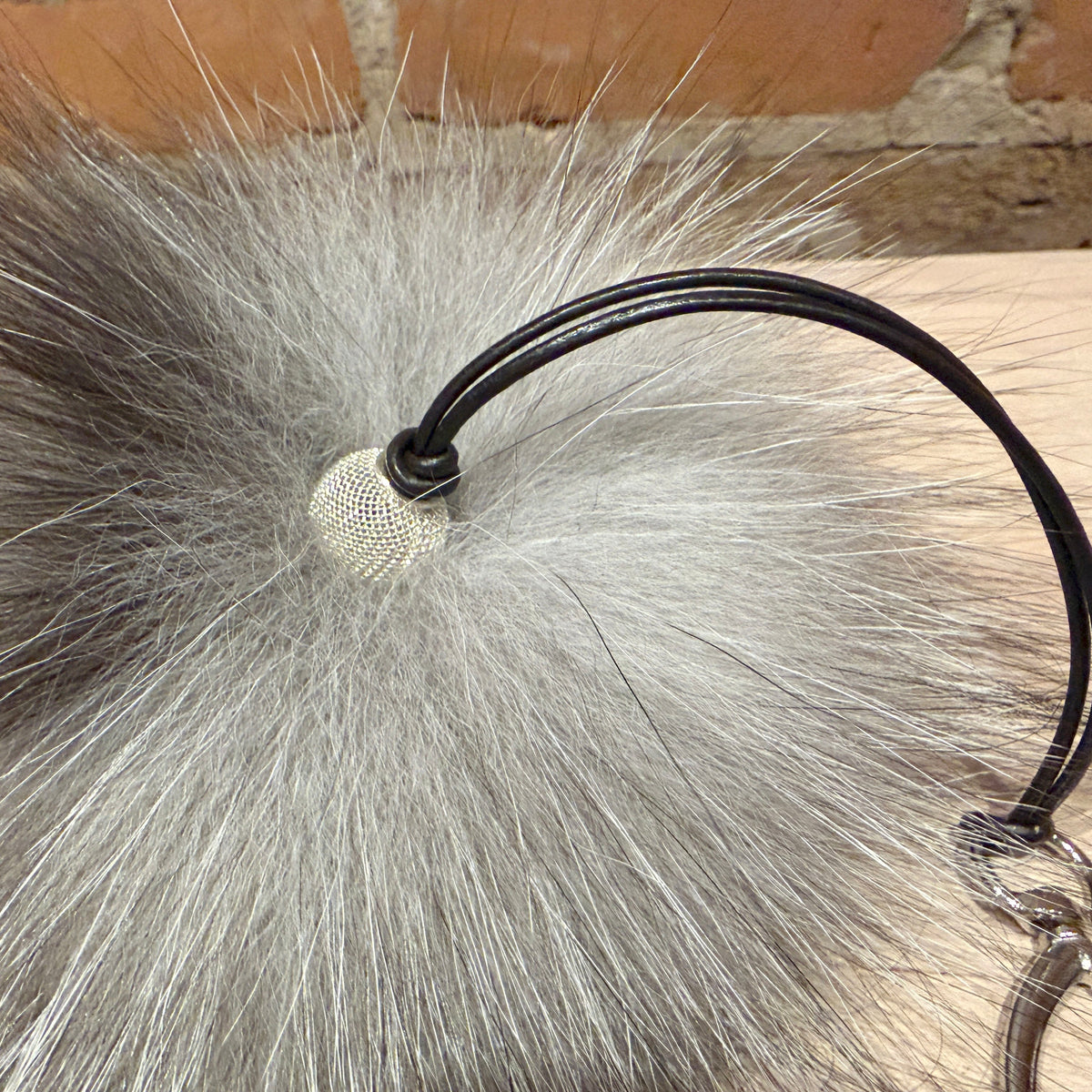 A close-up of a fluffy faux-fur pom-pom purse pom accessory in silver-gray and black, attached to a black elastic loop with a metal clasp, resting on a light wood surface with a brick wall in the background.
