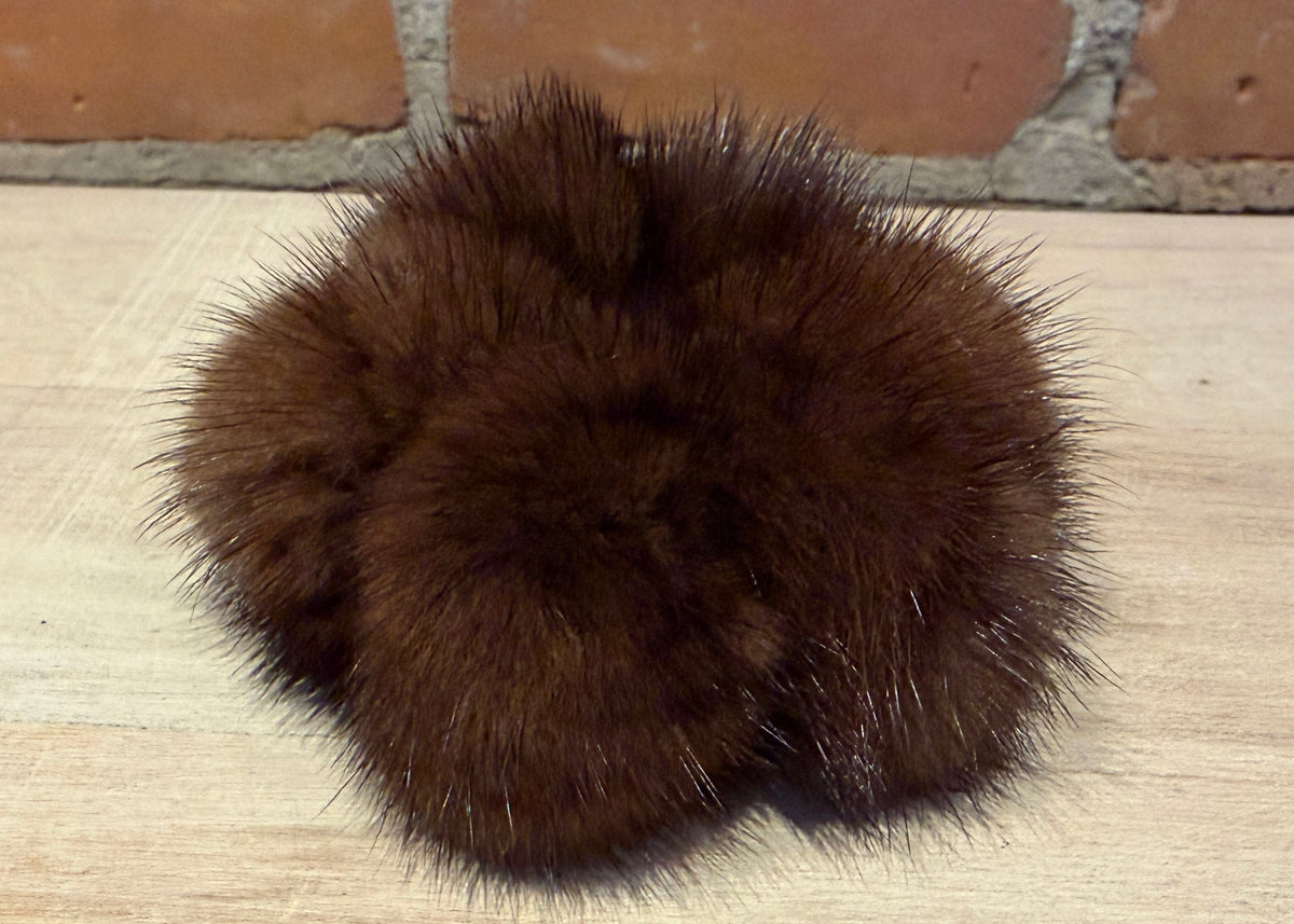 A dark brown fluffy mink fur pom-pom accessory resting on a light wood surface with a brick wall background.