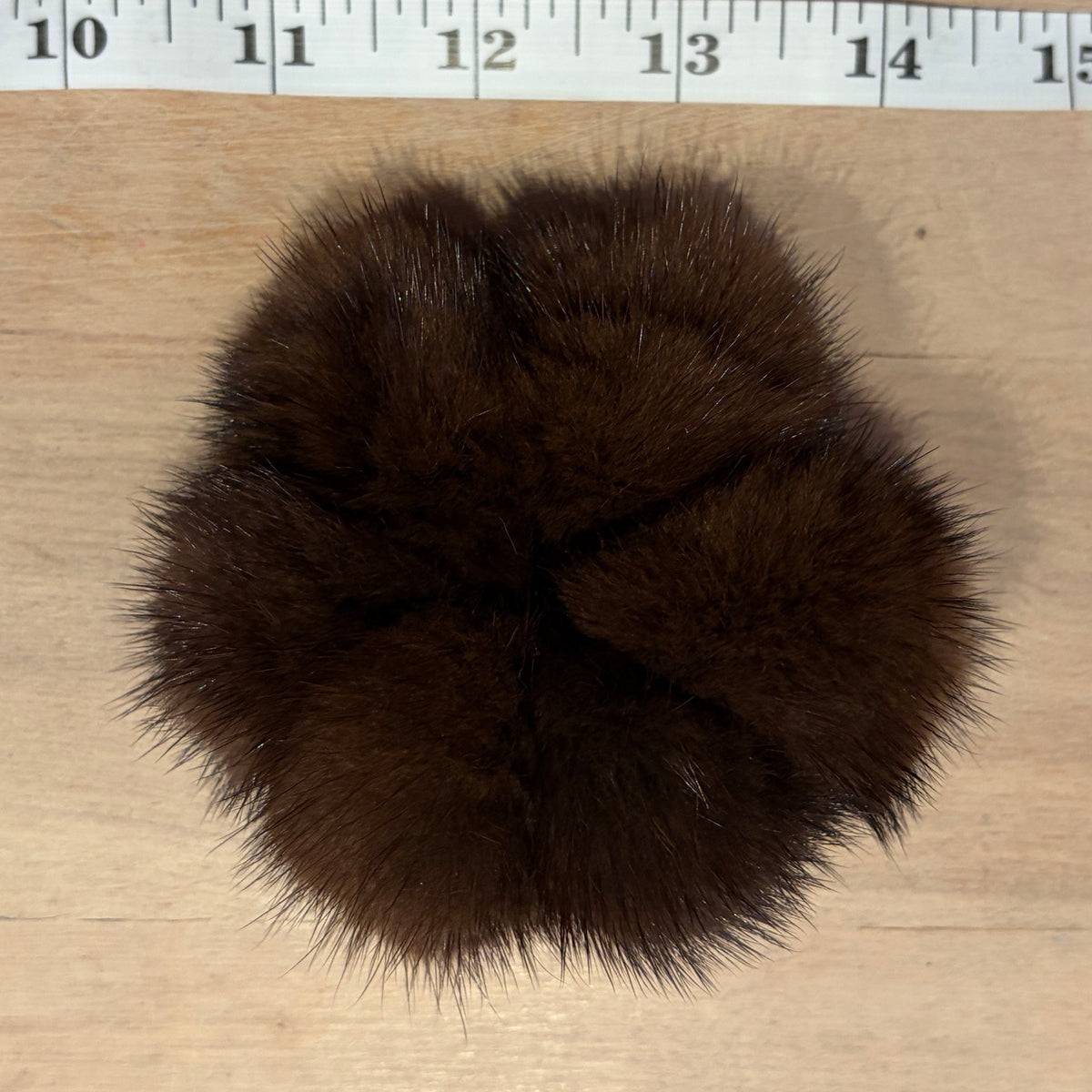 A dark brown mink fur pom-pom accessory, likely a hair scrunchie or decorative pom-pom, shown on a light wood surface with a measuring tape ruler at the top for scale.