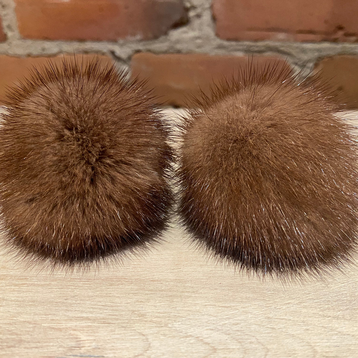 Two honey brown mink fur pom-poms with long, spiky tufts, displayed side-by-side on a light wood surface with a brick wall background.
