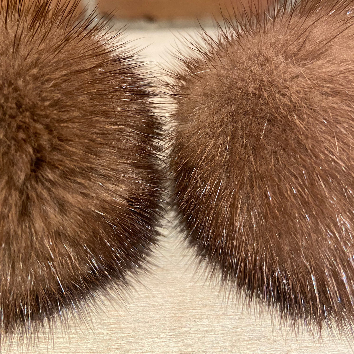 Two honey brown mink fur pom-poms with dark tips, shown close-up against a light wood surface and a brick wall background.