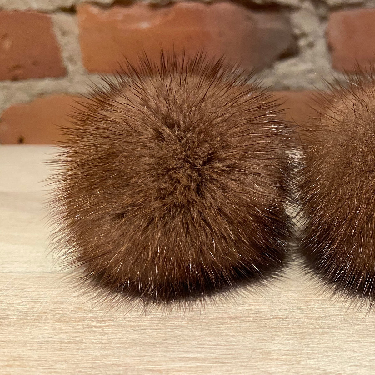 Two honey brown mink fur pom-poms with dark tips, displayed side-by-side on a light wood surface with a brick wall background.