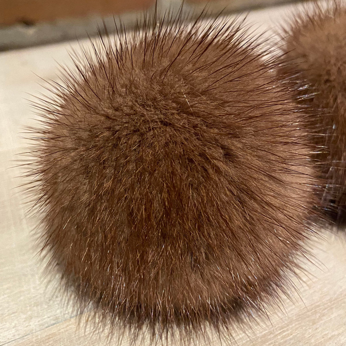 A close-up of a brown furry spherical object with dense bristles, shown on a light wood surface with another similar item partially visible at the right edge.