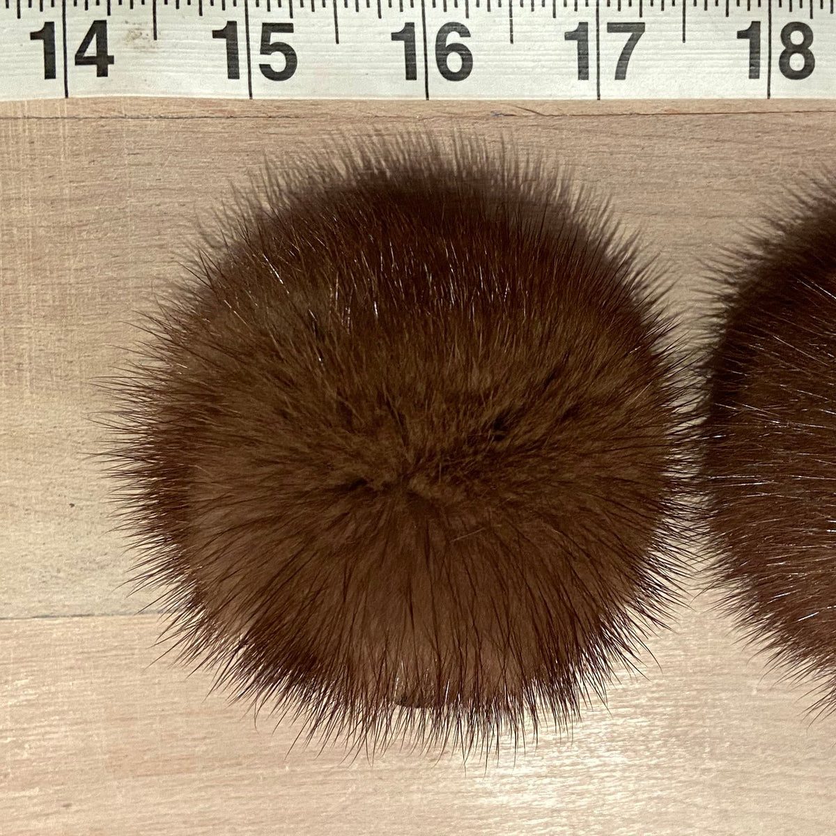 Two round honey brown mink fur pom-poms displayed side-by-side on a light wood surface with a measuring tape ruler at the top to indicate size.