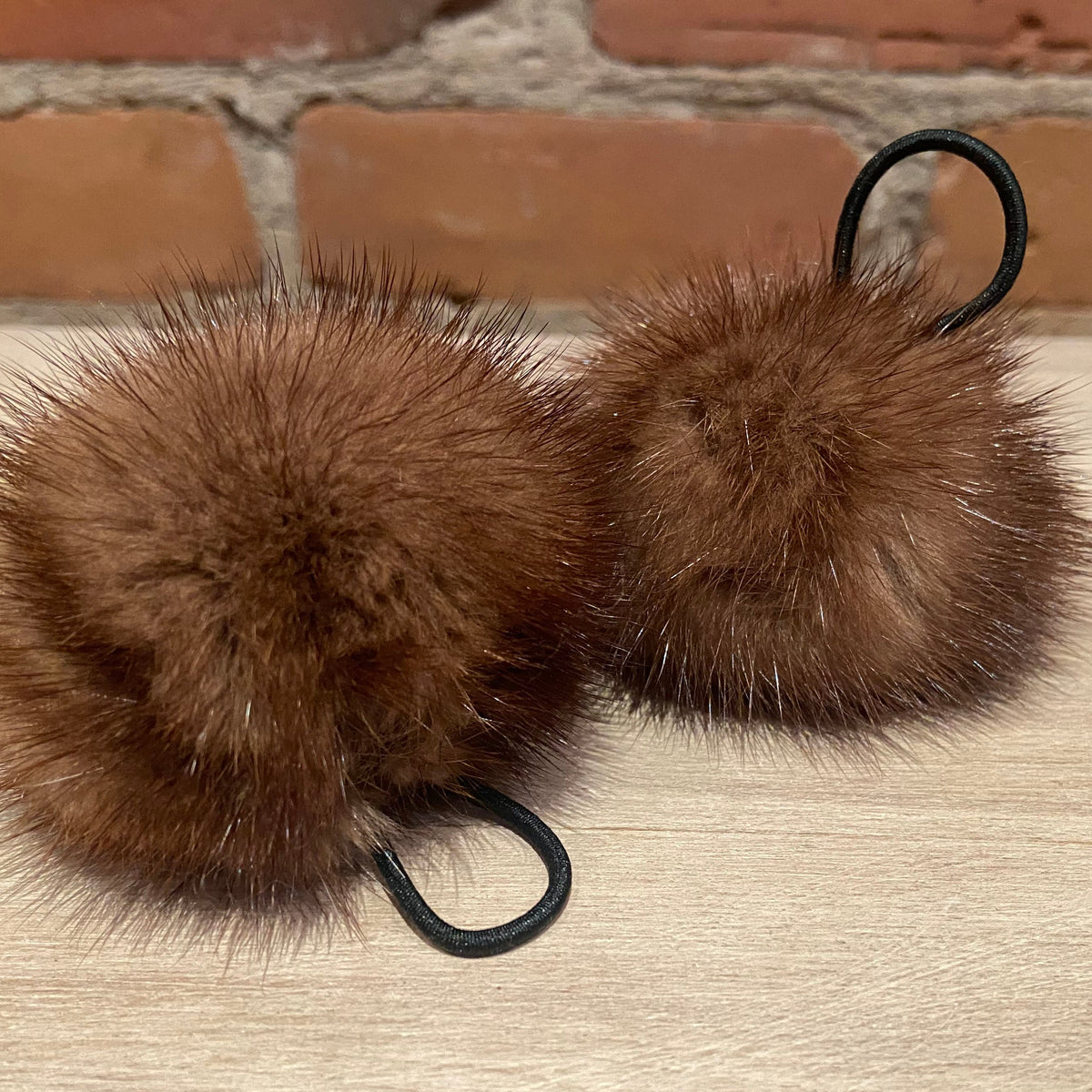 Two honey brown mink fur pom-poms with black elastic loop attachments  resting on a light wood surface with a brick wall background.