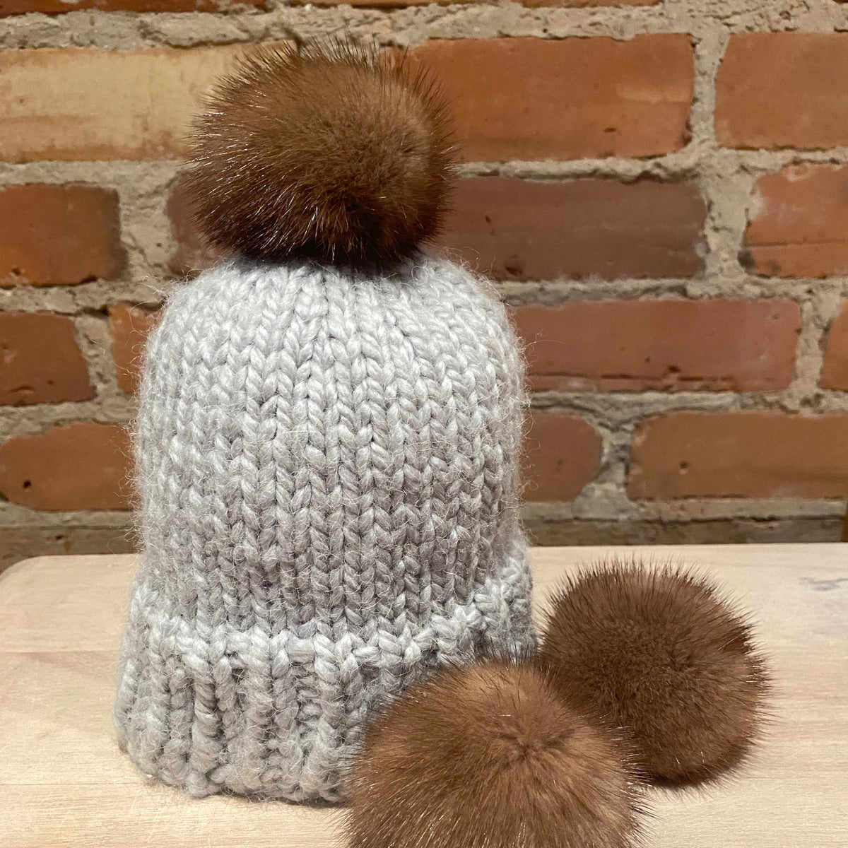 A small light gray chunky knit beanie with a small honey brown mink fur pom-pom on top and two matching brown pom-poms resting in front, set on a light wood surface against a brick wall background.