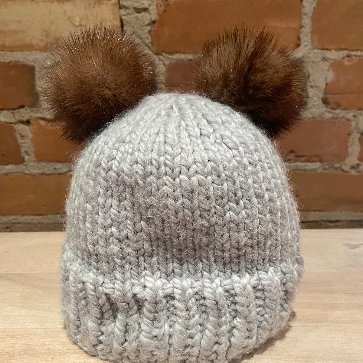 A light gray chunky knit beanie hat with two honey brown mink-fur pom-poms on top, displayed on a wooden surface against a brick wall background.