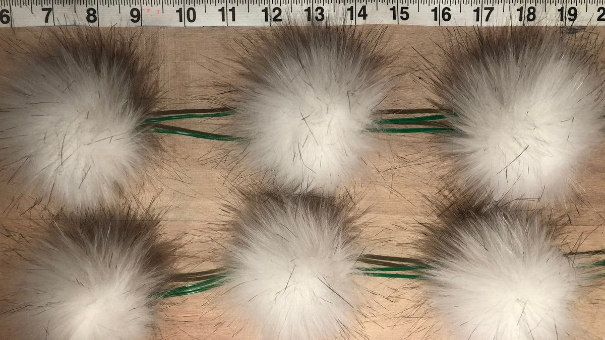 Six white fluffy faux-fur pom-poms with green wire handles arranged on a wooden surface below a measuring tape ruler.