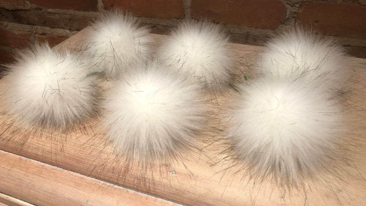 Six fluffy white faux fur pom-poms with long dark-tipped fibers, arranged on a light wood surface against a brick wall background.