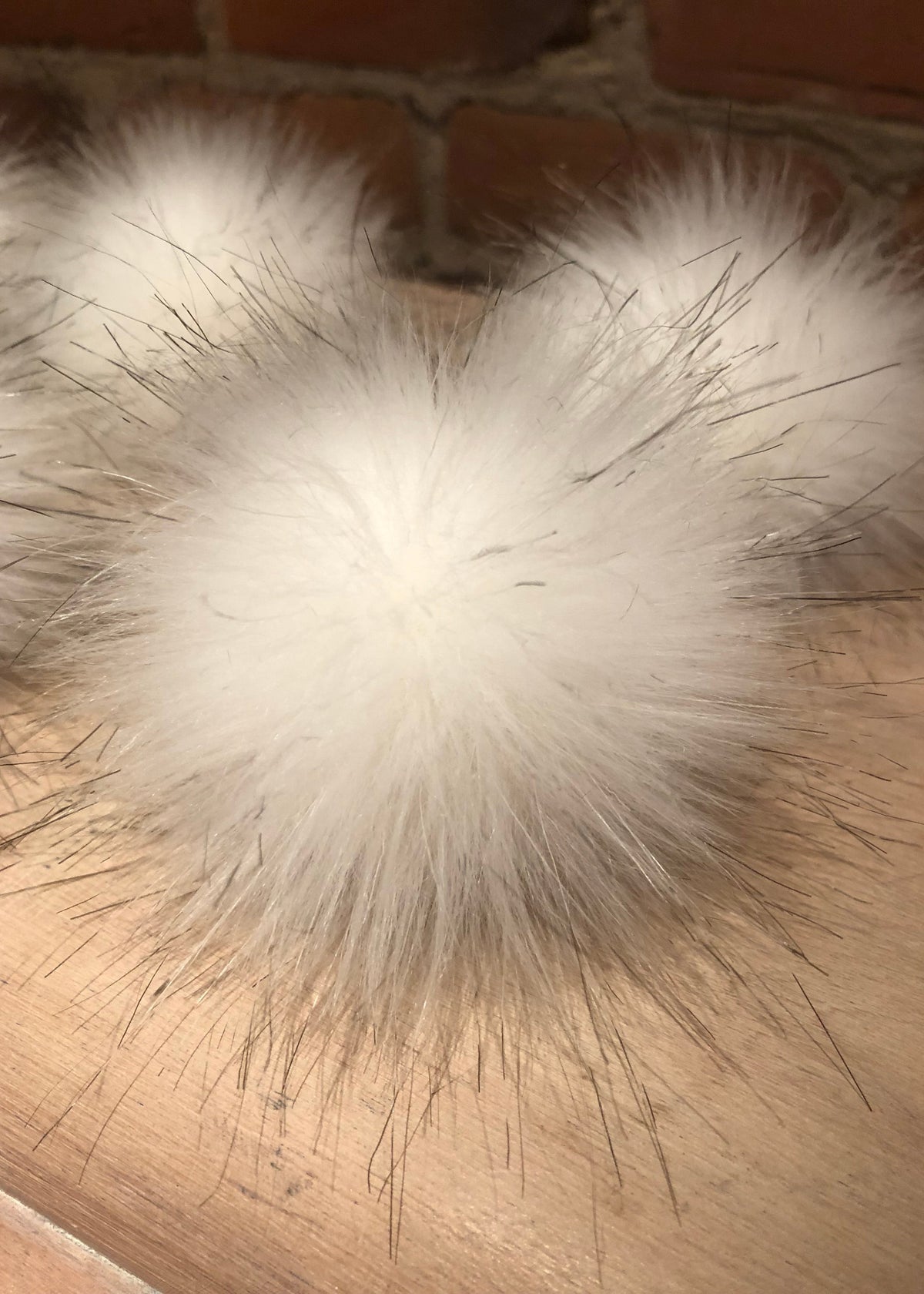 A close-up of a fluffy white faux fur pom-pom with dark guard hairs, resting on a light wood surface with a brick wall in the background.