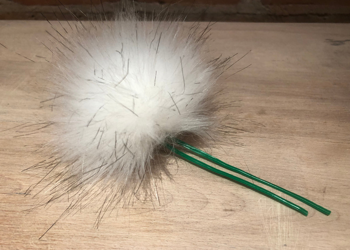 A white faux fur pom-pom hair accessory with green plastic wire stems, resting on a light wood tabletop.