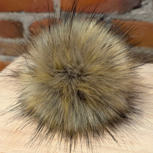 A fluffy faux fur pom-pom ball in a natural blend of beige, brown, and black, resting on a light wood surface with a blurred brick wall background.