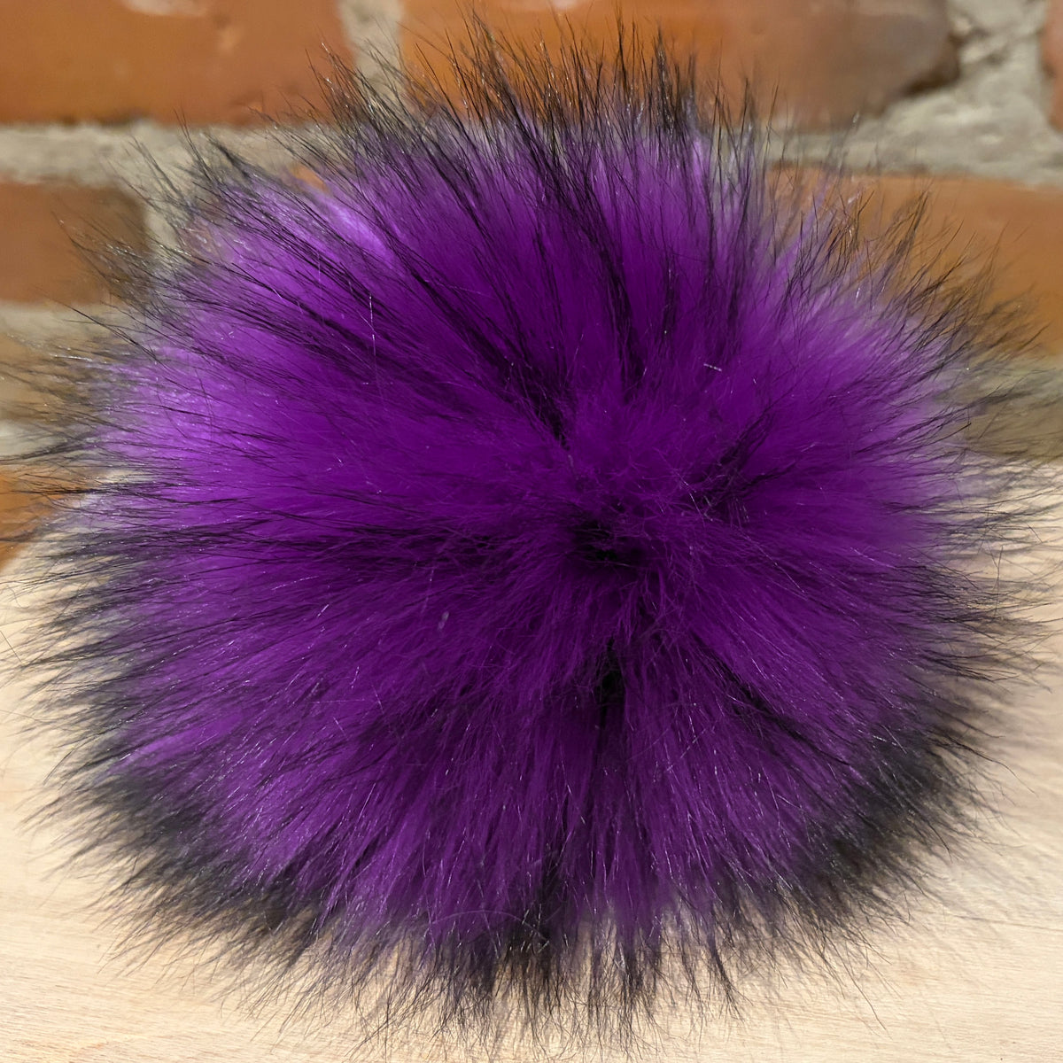 A round, fluffy pom-pom accessory in vibrant purple with black-tipped fibers around the edges, resting on a light wood surface with a blurred brick wall background.