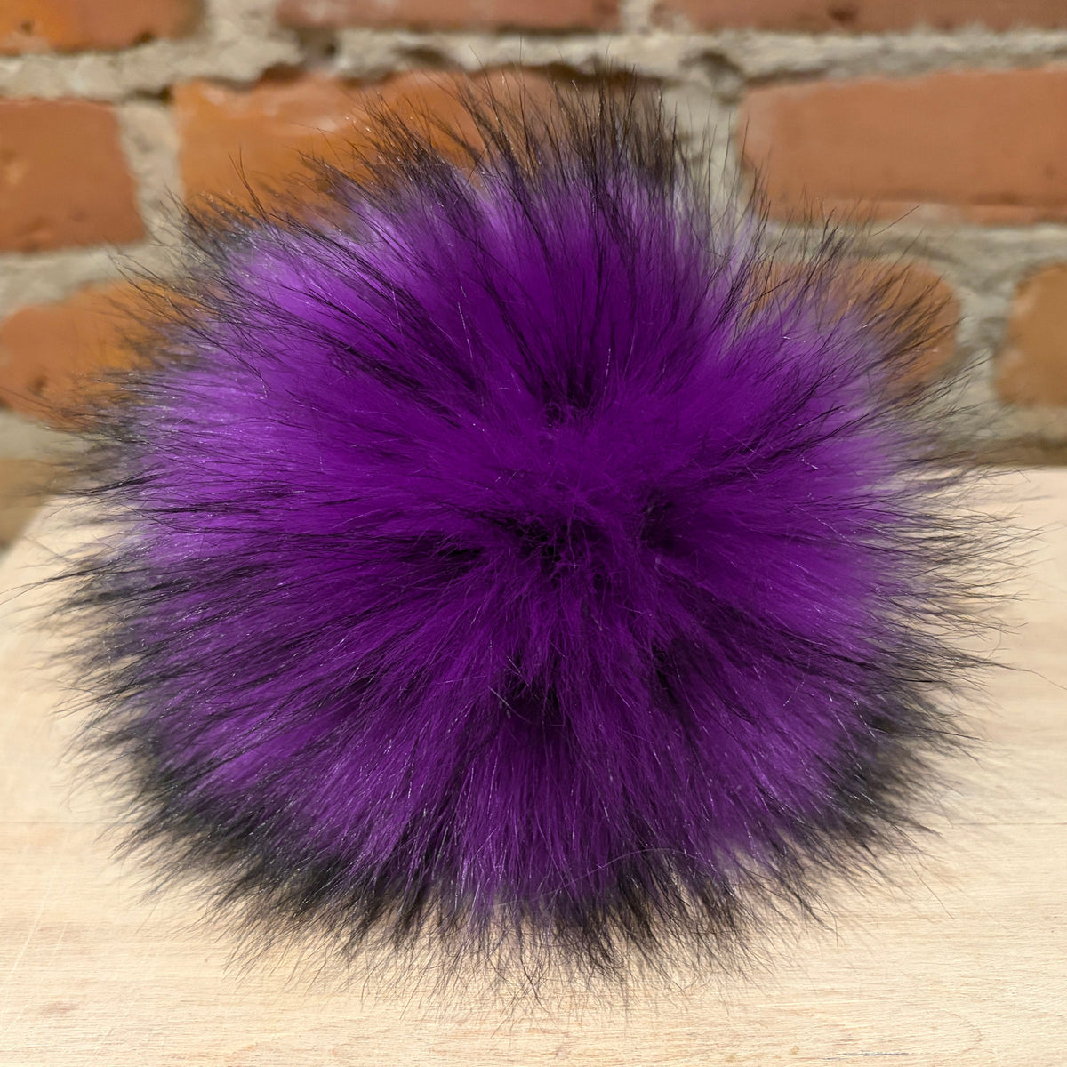 A fluffy pom-pom accessory in vibrant purple with black-tipped fur strands, resting on a light wood surface against a brick wall background.