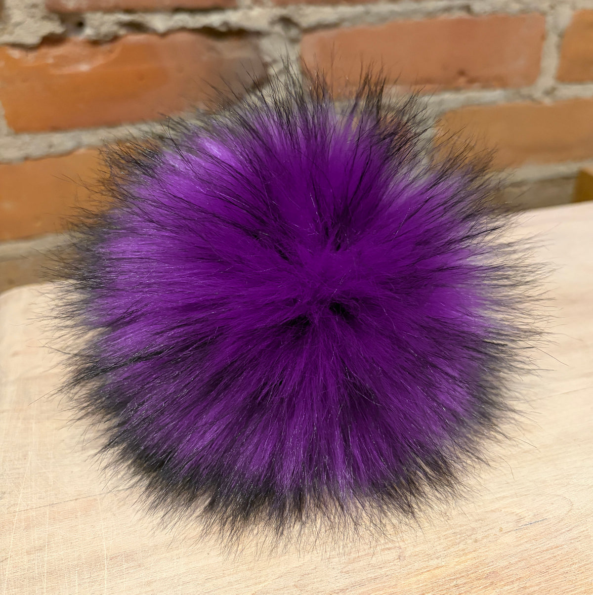 A fluffy pom-pom accessory in vibrant purple with black tips, resting on a light wood surface with a brick wall background.