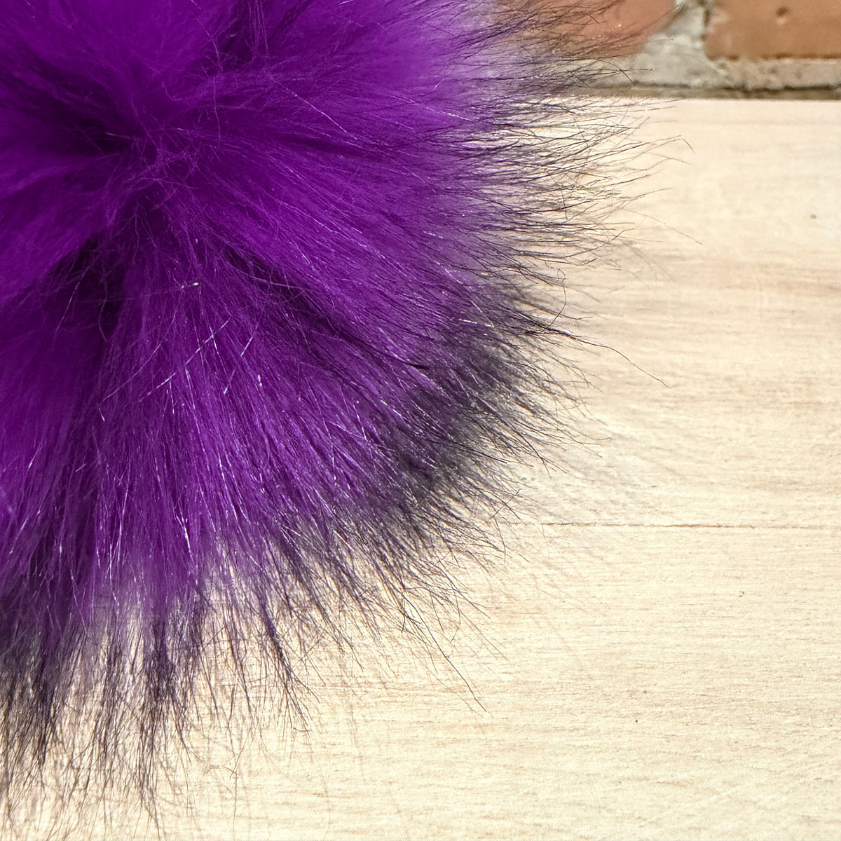 A close-up of a fluffy purple faux fur pom-pom resting on a light wood surface with a brick wall in the background.