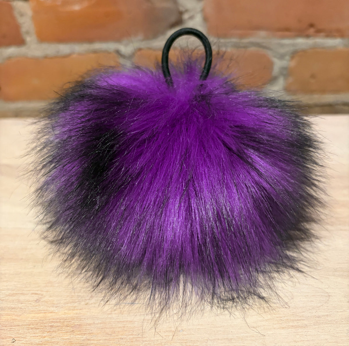 A fluffy faux fur pom-pom keychain or bag charm in vibrant purple fading to black, attached to a black metal ring, resting on a light wood surface with a brick wall background.
